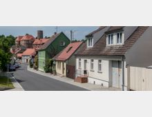 Straßenszene in einer ländlichen deutschen Kleinstadt mit Reihen von Häusern mit roten und grauen Ziegeldächern, darunter ein grünes Gebäude. Im Hintergrund ist eine Burg mit Rundturm und roten Dächern zu sehen, umgeben von grünen Bäumen.