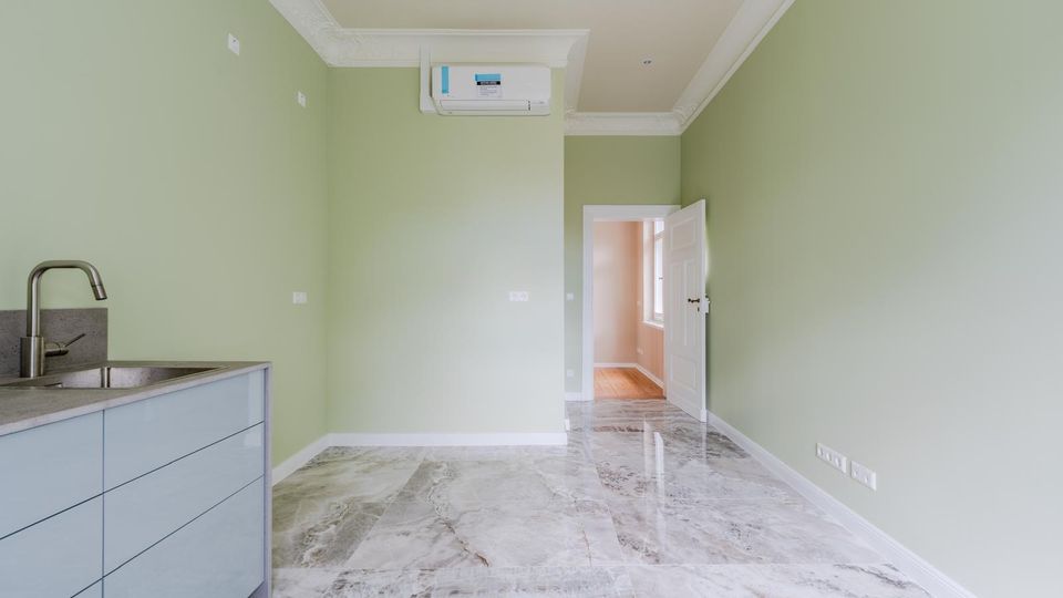 Leerer, schmaler Küchen- oder Hauswirtschaftsraum mit hellgrünen Wänden, glänzendem Marmorfliesenboden und einer kleinen Küchenzeile mit Spüle links. An der Stirnwand ist eine Klimaanlage montiert; eine offene Tür führt in einen angrenzenden Flur mit Fensterlicht.
