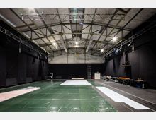 Großer, leerer Veranstaltungsraum mit hohen Decken und sichtbaren Stahlträgern. Der Boden ist teilweise mit grünen und weißen Folien abgedeckt, schwarze Wände und technische Installationen wie Lichttraversen und Kabel markieren den Raum.