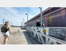 Eine erhöhte, breite Fußgängerbrücke führt zu einem modernen S-Bahnhof mit der Aufschrift „Warschauer Straße“. Der Bereich ist mit Laternen und Geländern versehen, die von Graffiti bedeckt sind, während eine Person mit Rucksack auf der Brücke entlanggeht.