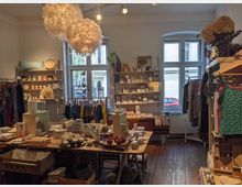 Ein kleiner Laden mit Holzboden und dekorativen Elementen. Es gibt Regale mit Keramik, Kleidung auf Kleiderbügeln und einen Tisch mit verschiedenen Geschenkartikeln, darunter Kerzen und kleine Dekorationen.