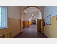 Langer Schulflur mit hohen Rundbögen, glattem Boden und einer Reihe kugelförmiger Deckenlampen, der zu einer Doppeltür am Ende führt. Links sind große Fenster und ein Heizkörper, rechts ein schwarzes Brett und ein Feuerlöscher; entlang des Flurs stehen orange-gelbe Schließfächer.