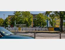 Eine moderne Schul- oder Bürogebäudeanlage mit gelb-blauen Fassaden, umgeben von Bäumen. Im Vordergrund befinden sich eine Bushaltestelle mit einer Glasüberdachung und eine mit Graffiti bemalte Mauer.