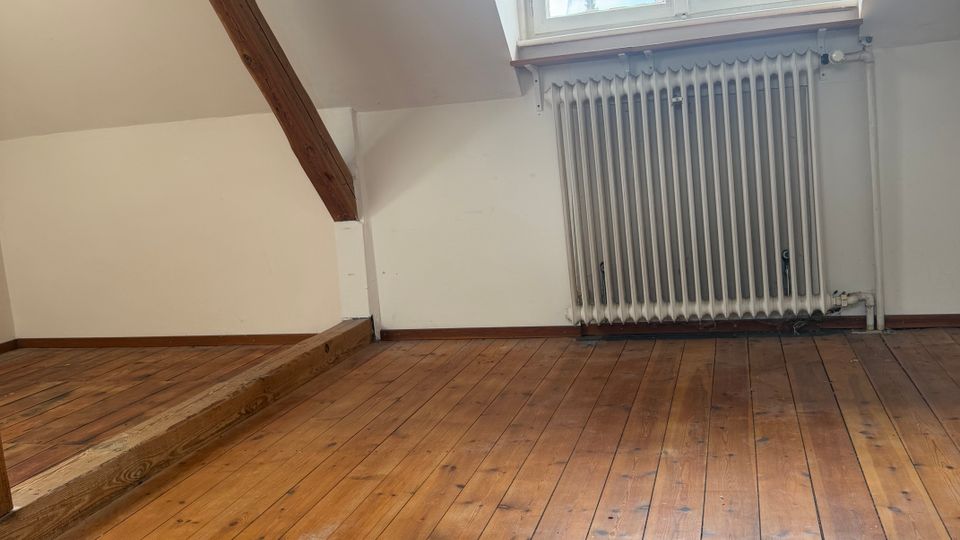 Leerer Dachraum mit schrägen weißen Wänden, sichtbaren Holzbalken und breiten Dielen-Holzboden. An der Stirnseite befindet sich ein kleines Dachfenster mit Heizkörper darunter.