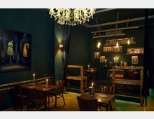 Gemütliches Café oder Restaurant mit dunklen grünen Wänden, einem Kronleuchter und Holzmöbeln. Im Hintergrund sind Bücherregale, Kerzenständer und eine dekorative Kaminattrappe zu sehen, die für eine stimmungsvolle Atmosphäre sorgen.