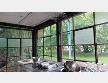 Ein Raum mit großen Fensterfronten aus Metallrahmen, die den Blick auf eine grüne Parklandschaft freigeben. Im Vordergrund stehen Küchengeräte, darunter Schüsseln, Tabletts und Gläser, auf Edelstahloberflächen.