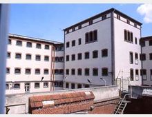 Mehrstöckige Gebäudekomplexe mit weißen Fassaden und symmetrischen Fenstern, umgeben von hohen Sicherheitsmauern; im Vordergrund Ziegeldächer und Gitter. Der Himmel ist klar und blau.