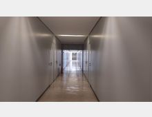 Langer, gerader Innenflur mit hellgrauen Wänden und mehreren Türen auf beiden Seiten; die glänzenden Bodenfliesen reflektieren das Licht, und an der Decke sind Rasterplatten mit integrierter Beleuchtung zu sehen. Am Ende des Flurs führt eine verglaste Tür in einen weiteren Bereich.