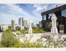 Dachterrasse mit Sitzbereichen, Klappstühlen und geschlossenen Sonnenschirmen, umgeben von Pflanzen und Blumen; im Hintergrund eine Stadtlandschaft mit modernen Gebäuden und dem Berliner Fernsehturm.