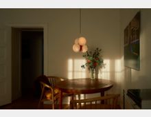 Esszimmer mit einem runden Holztisch und zwei Holzstühlen, beleuchtet von warmem Sonnenlicht. Über dem Tisch hängt eine moderne Pendelleuchte mit kugelförmigen Lampen, daneben steht eine Vase mit Blumen auf dem Tisch.