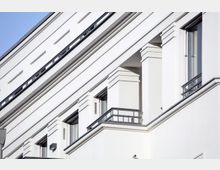 Fassade eines modernen weißen Gebäudeabschnitts mit klaren Linien, verzierten Balkongeländern aus Metall und rechteckigen Fenstern, teilweise umrahmt von großen dekorativen Pfeilern. Im Vordergrund sind Balkone sichtbar.