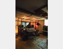 Tonstudio mit einem hölzernen Mischpult-Tisch, zwei großen Lautsprechern, einem Mischpult und weiterer Studioausrüstung. Der Raum hat Holzelemente an Decke und Wänden, sanfte Beleuchtung und einen Teppich auf einem Holzboden.