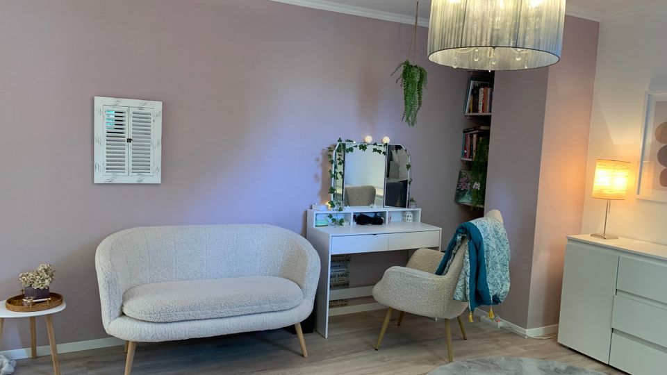 Helles Wohnzimmer mit rosafarbenen Wänden und grauem Laminatboden, eingerichtet mit einem cremefarbenen Sofa, einem Schminktisch mit Spiegel und passendem Sessel sowie einem runden Teppich. Rechts stehen ein Sideboard mit Stehlampe und ein Regal in einer Nische, darüber hängt eine große Deckenleuchte mit Stoffschirm.