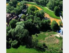 Luftaufnahme einer grünen Landschaft mit einem kleinen Fluss mit orange-braunem Wasser, der sich durch das Gelände schlängelt. Links im Bild stehen mehrere Gebäude umgeben von Bäumen, rechts eine Brücke, die den Fluss überquert.