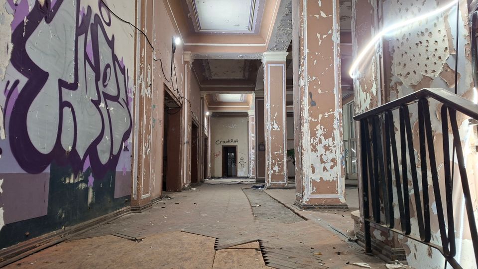 Verlassener Innenflur eines alten Gebäudes mit hohen Decken, Oberlichtern und Säulen; die Wände sind stark abgeblättert und teilweise mit Graffiti bedeckt. Im Vordergrund liegen beschädigte Bodenplatten und Schutt, rechts ist ein Treppengeländer zu sehen.
