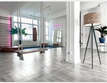 Großzügiger, moderner Raum mit hellen Holzböden und großen Fenstern, die viel Tageslicht hereinlassen. Im Vordergrund eine Holzschaukel an Seilen, ein Boxsack, Pflanzen in Töpfen und eine Stehleuchte; im Hintergrund Sportgeräte wie eine Kletterwand und eine blaue Bodenmatte.