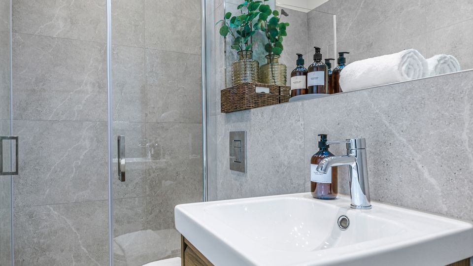 Modernes Badezimmer mit grauen Steinfliesen, bodengleicher Dusche mit Glasabtrennung und Waschtisch mit weißem Aufsatzbecken auf Holzunterschrank. Integrierte Ablage mit Handtüchern und Pflegeprodukten sowie Decken-Spots und Schienenbeleuchtung.