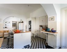 Helle Küche mit Retro-Elementen, ausgestattet mit einer weißen Küchenzeile, einem freistehenden Küchentresen mit Holzoberfläche und schwarz-weißen Schachbrettfliesen. Im Hintergrund sind ein Rundbogen, Schränke, Regale und Wandbilder sichtbar.