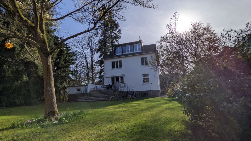 Freistehendes, weißes Wohnhaus mit mehreren Stockwerken und Dachgauben, über eine breite Treppe von einem großen, grünen Garten aus erreichbar. Umgeben von hohen Bäumen und Sträuchern unter hellem Himmel mit tief stehender Sonne.
