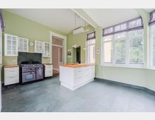 Helle Wohnküche mit grün gestrichenen Wänden, weißen Schränken mit Glasfronten und einer Kochinsel mit Holzarbeitsplatte. Große Fenster mit Raffrollos, dunkler Fliesenboden und freistehender Herd vor dunkler Rückwand prägen den Raum.