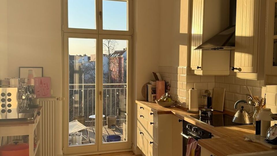 Sunlit residential kitchen with white shaker-style cabinets, wooden countertops, a white tile backsplash, and a black range hood over the stove. A tall glass door with divided panes opens to a small balcony, and a black pendant light hangs above wide wooden floorboards.