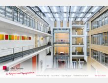 Moderner Innenraum eines Konferenz- und Tagungszentrums mit einer zentralen, offenen Galerie und mehreren Etagen. Glasgeländer, Holztüren, helle Wände, farbige Wandbilder sowie ein Oberlicht mit moderner Beleuchtung prägen die Architektur.