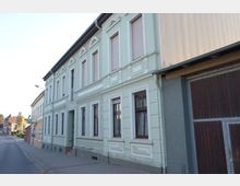 Straßenszene mit einem hellgrünen, zweigeschossigen Wohngebäude im Vordergrund, das mit verzierten Fensterrahmen und Rollläden gestaltet ist. Rechts schließt ein graues Gebäude mit einer großen Holztoröffnung an, während die Straße und weitere Häuser in die Ferne führen.