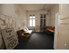 Ein einfacher, vernachlässigter Raum mit abblätternder Wandfarbe, zwei Fenstern mit weißem Holzrahmen, einem grauen Sofa und einem braunen Sessel. Auf der rechten Seite befinden sich ein Tisch mit Stühlen sowie ein Einkaufswagen; auf dem Boden liegt ein dunkler Teppichboden. Ein großes Schild lehnt an der linken Wand.