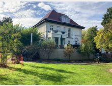 Zweistöckiges Wohnhaus mit hellgelber Fassade und dunkelrotem Walmdach, umgeben von einem Garten mit grüner Rasenfläche, mehreren Bäumen und Sträuchern. Im Vordergrund ist ein kleiner Weg und Gartenwerkzeug sichtbar, während die Terrasse des Hauses mit Geländer und Dekorationen ausgestattet ist.