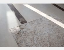 Teilausschnitt eines Raumes mit hellem, glatten Boden, der von Sonnenlicht durchflutet wird. Am unteren Bildrand ist eine Ecke eines beigefarbenen Teppichs mit ornamentalem Muster sichtbar, neben einem dunklen Streifen im Bodenbelag.