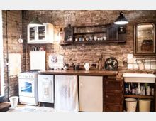 Rustikale Küche mit freiliegenden Ziegelwänden, offener Holzregalaufbewahrung und einer Arbeitsplatte aus Holz. Zu sehen sind ein weißer Gasherd, eine Spüle, ein Spiegel mit goldfarbenem Rahmen sowie verschiedene Geschirr- und Dekorationsgegenstände.