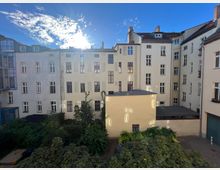 Hinterhof mit einem Mehrfamilienhaus in geschlossener Bauweise. Die Fassade besteht aus hell verputzten Wänden mit zahlreichen Fenstern, im Vordergrund sind Sträucher und Bäume sowie ein kleiner, begrünter Bereich zu sehen.