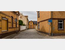 Schmale gepflasterte Wohnstraße, gesäumt von Häusern mit gelben Ziegelsteinen und braunen Verzierungen. Im Hintergrund steht ein kleines, ebenfalls gelbes Wohngebäude mit Spitzdach, und ein Straßenschild weist auf eine Verkehrsberuhigte Zone hin.