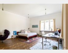 Helles Schlafzimmer mit Holzboden, einem Bett auf einem gemusterten Teppich, einem Schreibtisch mit Bürostuhl auf einem weiteren Teppich und einem großen Fenster mit beigefarbenen Vorhängen. Im Raum stehen zudem ein Sessel und ein kleiner Beistelltisch.