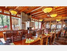 Rustikales Restaurant mit Holzmöbeln und Holzbalkendecke, großen Fenstern, die Tageslicht hereinlassen, sowie dekorativen Hängelampen. Ein Kaminofen ist in eine gemauerte Säule integriert, und der Raum bietet Sicht auf eine waldähnliche Außenlandschaft.