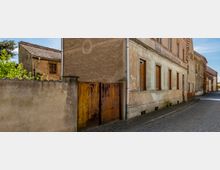 Straßenszene mit alten Wohnhäusern in einem verfallenen Zustand, mit bröckelnden Fassaden und geschlossenen Fensterläden; links ein rostiges Metalltor neben einer niedrigen Betonmauer, im Hintergrund Bäume und blauer Himmel.