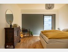 Schlafzimmer mit Holzboden und modernen Elementen. Zu sehen sind ein Bett mit gelber Bettwäsche, eine antike Kommode mit einem ovalen Spiegel, eine Gitarre, ein geflochtener Stuhl, einige Pflanzen und eine Glastür, die auf einen Balkon führt.