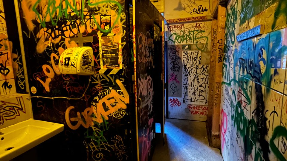 Schmaler, industriell wirkender Innenraum mit Betonboden, sichtbaren Lüftungsrohren an der Decke und warmem Kunstlicht. Wände, Türen und Trennwände sind dicht mit Graffiti und Stickern bedeckt; links ist ein kleines Waschbecken zu sehen.