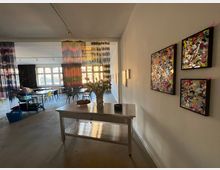 Ein heller Innenraum mit einer offenen Gestaltung, dekoriert mit bunten Kunstwerken an den Wänden und langen, farbigen Stoffvorhängen, die den Raum visuell trennen. Im Vordergrund steht ein Tisch mit einer Vase voller Blumen, während im Hintergrund Tische und Stühle vor großen Fenstern zu sehen sind.