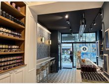 Innenraum eines stilvollen Cafés mit modernem Design: Links ein Regal mit Gläsern und Produkten, rechts eine Theke mit Gläsern und Snacks, daneben eine Sitzreihe mit hohen Stühlen. Durch die Glastür und Fenster im Hintergrund ist eine belebte Straße mit Fahrrädern und Passanten zu sehen.