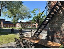 Innenhof eines historischen Backsteingebäudes mit großen Fenstern, umgeben von Bäumen. Im Vordergrund ein Holztisch mit Stühlen, eine Metalltreppe an der Wand und ein Schornstein im Hintergrund.