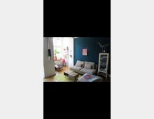 Wohnzimmer mit moderner Einrichtung, bestehend aus einem beigefarbenen Sofa, einem Holzcouchtisch und einem bunten Teppich. Eine dunkelblaue Wand mit einem kleinen Bild, daneben ein Glaskasten mit Geschirr, und helle Fensterfront mit Pflanzen im Hintergrund.