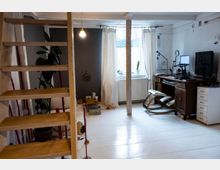 Heller Wohnraum mit weiß gestrichenen Holzböden, einer offenen Holztreppe auf der linken Seite und einem Arbeitsplatz auf der rechten Seite, bestehend aus einem dunklen Holzschreibtisch, Monitor und Schaukelstuhl. Im Hintergrund steht ein großes Fenster mit beigen Vorhängen, das den Raum beleuchtet.