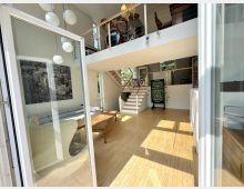 Heller, moderner Wohnraum mit hohen Decken, hellem Holzboden und großen Glasschiebetüren, die viel Tageslicht hereinlassen. Eine offene Treppe führt zu einer Galerie mit Geländer, auf der ein Schlagzeug steht; im Erdgeschoss sind Sitzbereich und ein Tischkicker zu sehen.