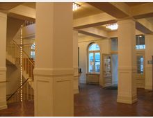 Heller Innenraum eines historischen Gebäudes mit weißen Wänden, hohen Pfeilern und Parkettboden. Im Hintergrund befinden sich eine Treppe mit Geländer, große Fenster mit Rundbogen und Doppeltüren mit Glaseinsätzen.
