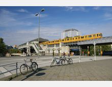 Eine erhöhte U-Bahn-Station mit einem vorbeifahrenden gelben Zug, flankiert von Treppenaufgängen und einer kleinen Statue. Im Vordergrund befindet sich ein Gehweg mit geparkten Fahrrädern, während im Hintergrund moderne Gebäude und eine teils eingehüllte Baustelle sichtbar sind.