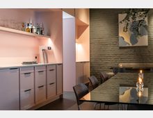 Moderne Innenaufnahme eines Raums mit einer Küchenzeile in Rosa mit Schränken, einer Theke und einer Wandhalterung für Glasflaschen. Rechts befindet sich ein Tisch mit schwarzer, glänzender Oberfläche, umgeben von Stühlen, und im Hintergrund eine Wand aus Sichtziegeln mit einem abstrakten Gemälde.