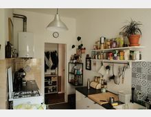 Kleine Wohnküche mit Gasherd, Arbeitsplatte und Spüle, über der offene Regale mit Vorratsgläsern, Gewürzen und hängenden Küchenutensilien montiert sind. Weiße Wände, eine große Hängelampe, gemusterte Fliesen an der Spüle sowie ein Durchgang zu einem Flur mit Garderobe prägen den Raum.