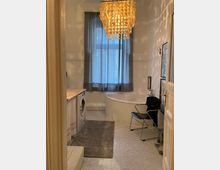 Badezimmer mit weißen Wänden und Bodenfliesen in Mosaikoptik, ausgestattet mit einer Eckbadewanne, Waschmaschine, grauem Teppich und einem freistehenden Stuhl. Ein dekorativer Kronleuchter aus runden Elementen hängt von der Decke, und ein großes Fenster mit grauem Vorhang befindet sich im Hintergrund.