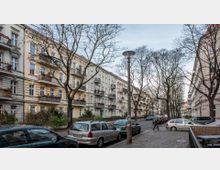 Straßenszene in einem Wohngebiet mit vierstöckigen Altbauhäusern, die pastellfarben gestrichen und mit Balkonen versehen sind. Die Straße ist gesäumt von kahlen Bäumen, parkenden Autos und einem gemusterten Gehweg aus Pflastersteinen.